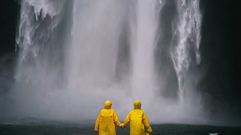 Zwei Menschen stehen mit knallgelben Regenmänteln händehaltend vor einem tosenden Wasserfall. 