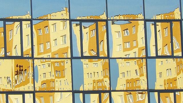 Häuserfront in hell- und dunkelgelb und hellblauer Himmel spiegeln sich in einer Fensterfront 