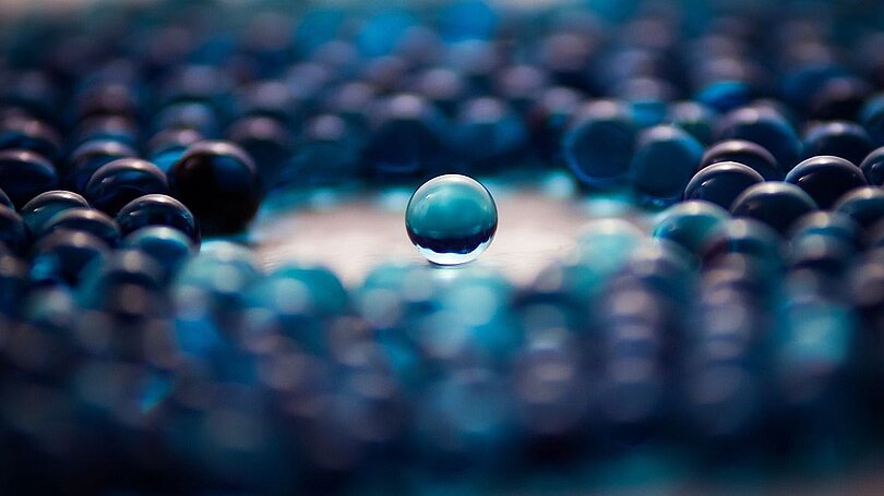 Am Tisch liegen blaue Glasmurmeln. In der Mitte ist ein Kreis frei und ganz in der Mitte liegt eine blaue Murmel allein.