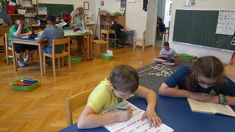 Ein Klassenraum: Ein Bub sitzt am Tisch und arbeitet in einem Schulbuch. Das Mädchen neben ihm lies ein Buch. Hinter ihnen sitzt am Boden ein Bub im Schneidersitz und arbeitet mit den Kärtchen vor sich. Am anderen Tisch sitzen drei Kinder und arbeiten. Eine Lehrerin steht an diesem Tisch und ist im Gespräch mit diesen Schüler*innen.