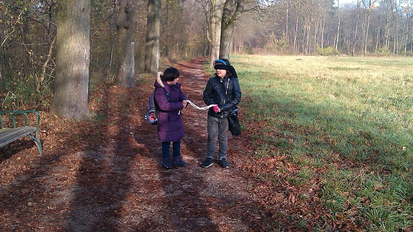 Zwei Kinder gehen nebeneinander in der Au. Beide haben jeweils ein Ende des gleichen Stück Seils in der Hand. Ein Kind führt, das andere folgt mit geschlossenen Augen. 