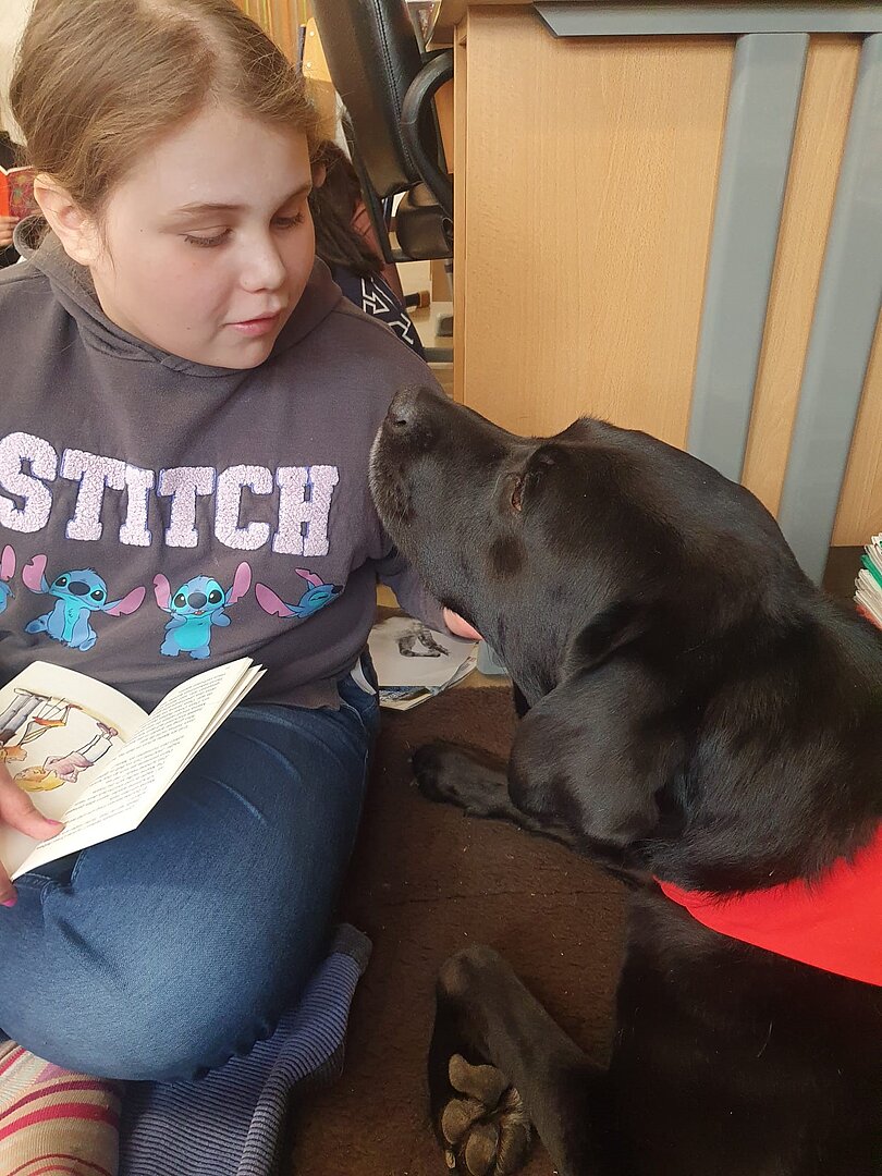 Originalfoto aus der Schule: Ein Mädchen sitzt im Schneidersitz am Boden, hat ein Buch auf ihren Beinen liegen und schaut liebevoll zum schwarzen Labrador neben ihr. 