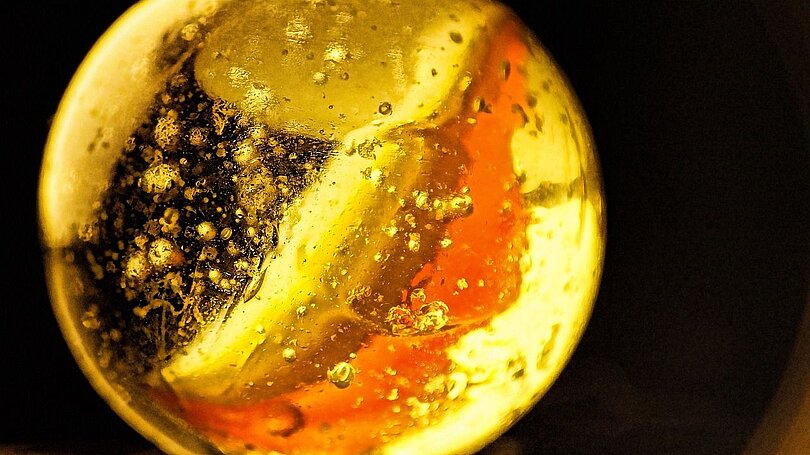 Ein weiße Glasmurmel mit einem orangen Streifen wird von hinten her angeleuchtet und leuchtet golden.