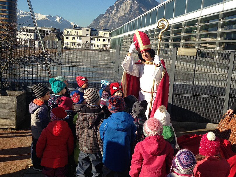 Ein Mann, gekleidet wie der Bischof Nikolaus steht im Freien. Er fasst sich an den Kopf bzw. an die Mytra und erzählt den Kindern darüber. Die Kinder stehen rundherum und hören zu. 
