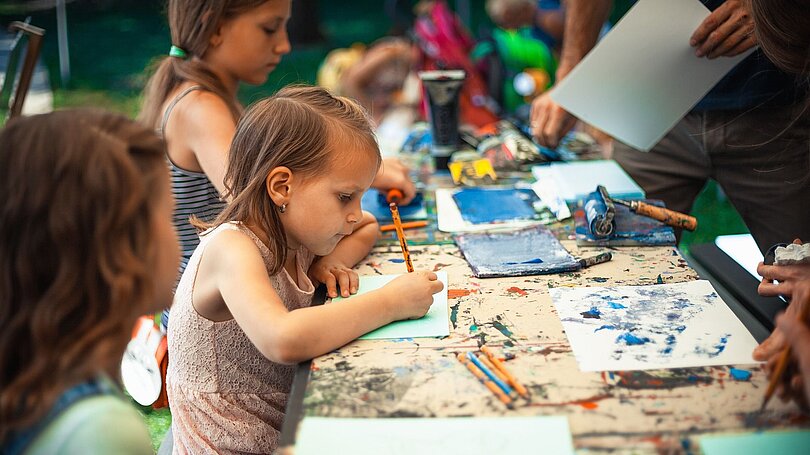 Drei Kinder sitzen an einer langen Mal-Tafel im Freien und sind ganz konzentriert bei der Arbeit. 